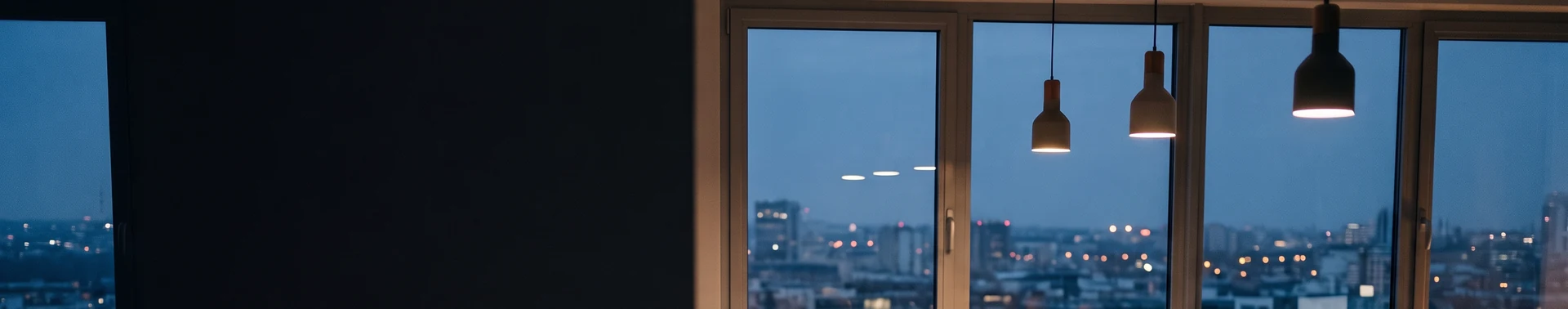 A dark office wall beside a row of windows at dusk, with pendant lamps and the lights of the city beyond